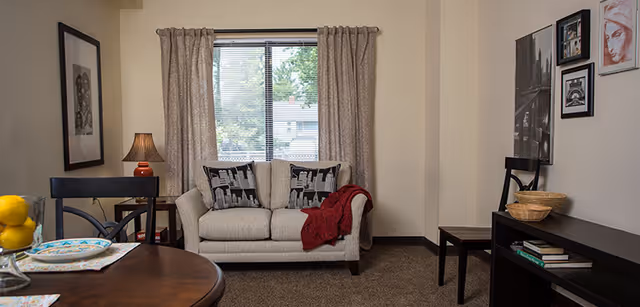 Cozy living room with a small loveseat, dining table, lamp, wall art, and a window with curtains.