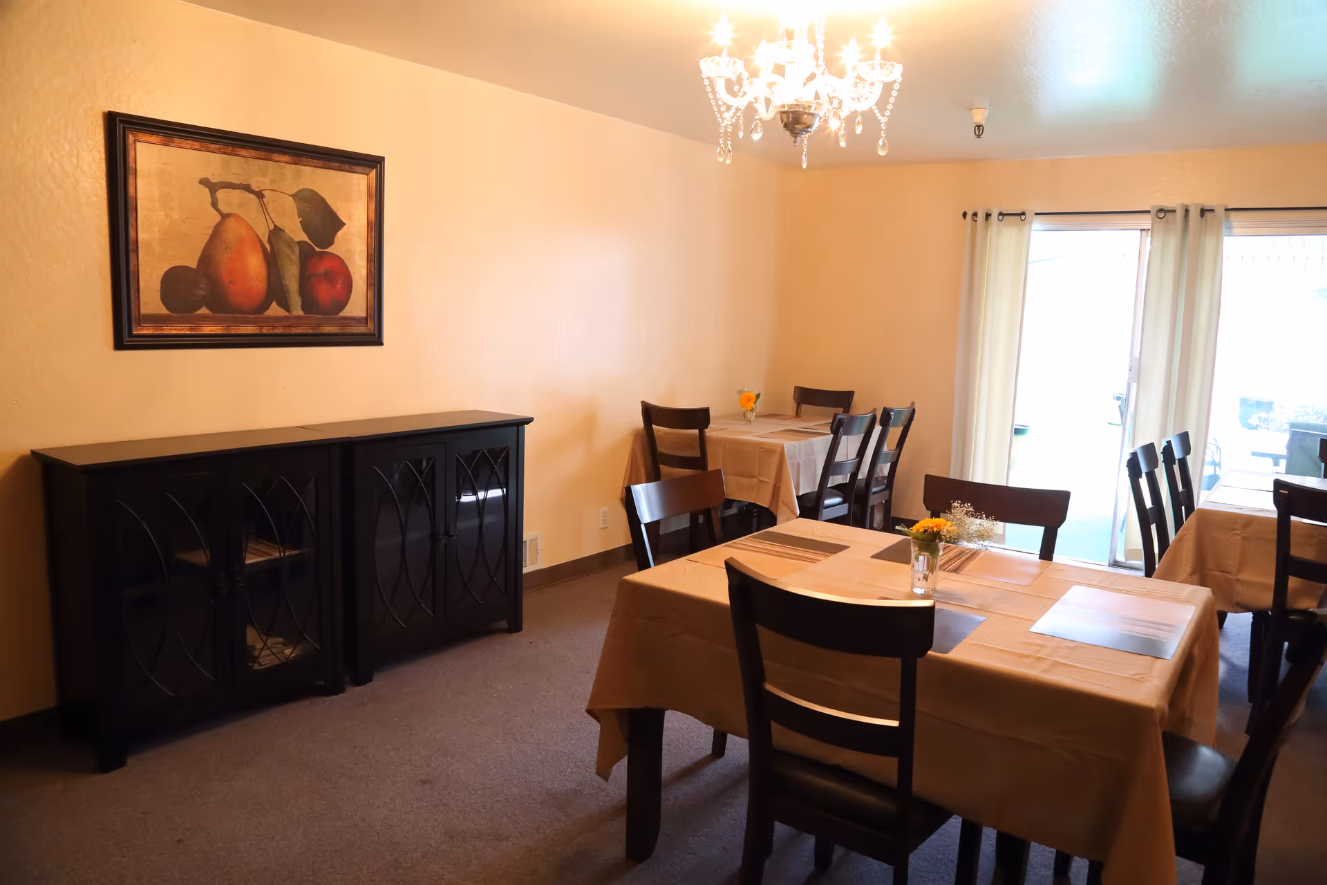 A dining room with several tables covered in beige tablecloths, each set with placemats and small flower arrangements. There are dark wooden chairs around the tables, a black cabinet against the wall, a framed painting of fruit above the cabinet, and a chandelier hanging from the ceiling. Sliding glass doors with light curtains lead to an outdoor area.