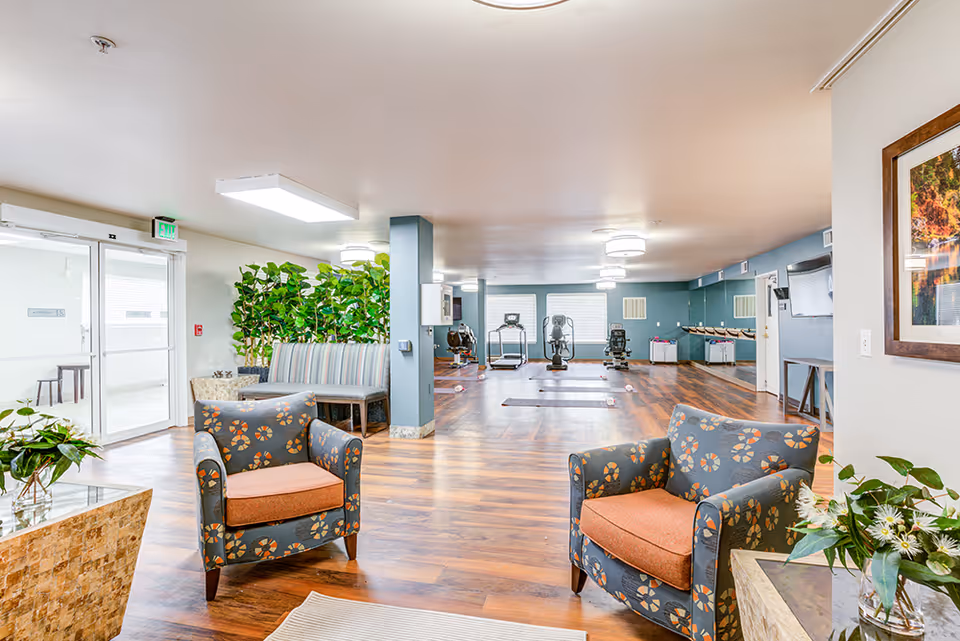 A spacious senior living common area with two patterned armchairs with orange cushions in the foreground, a glass coffee table with a plant, a bench with striped upholstery near a green leafy divider, and exercise equipment including treadmills and stationary bikes in the background. The room has wood flooring, light blue walls, and large windows letting in natural light.