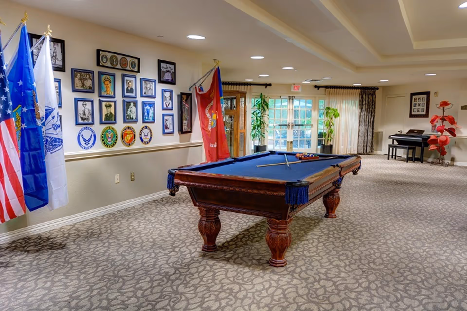A spacious room with a pool table in the center, surrounded by a patterned carpet. On the left wall, there are several framed pictures and military insignias, along with four flags. At the far end of the room, there are large windows with curtains, two potted plants, and a piano with a bench. The ceiling has recessed lighting and a tray ceiling design.