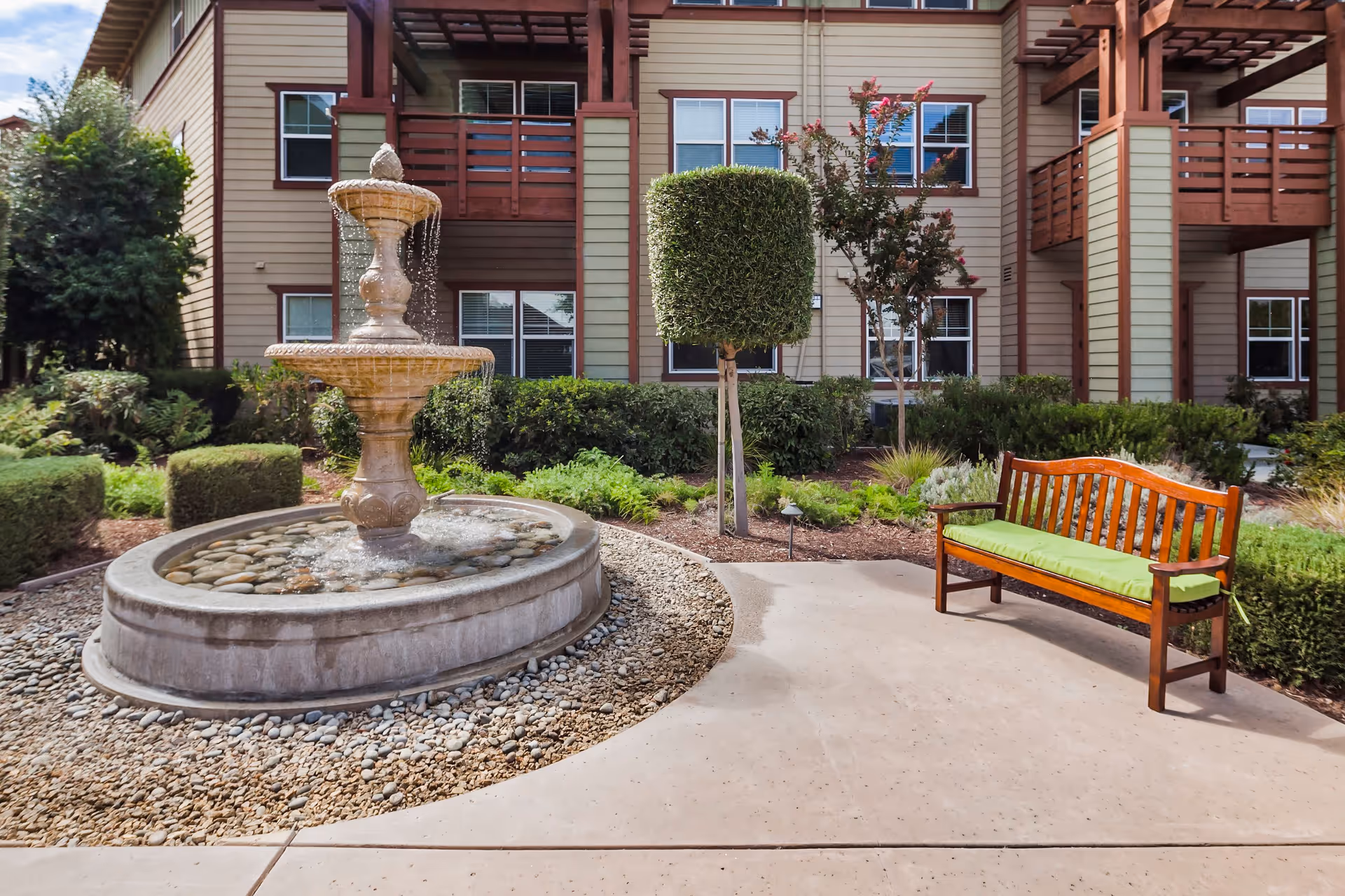 Outdoor courtyard area at Merrill Gardens at Gilroy featuring a stone water fountain surrounded by small rocks, a wooden bench with a green cushion, manicured bushes, and a multi-story building with balconies and windows in the background.