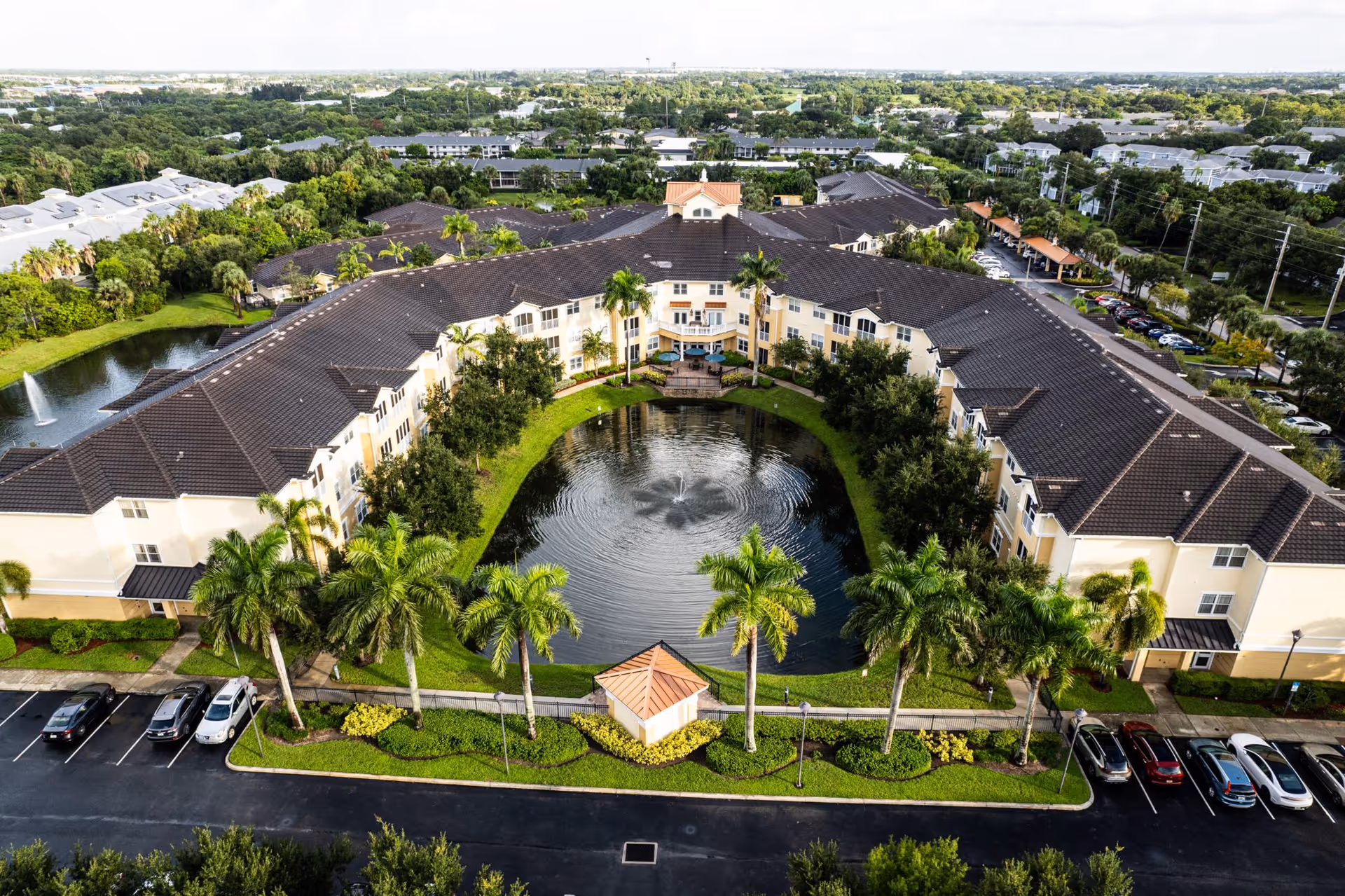 Aerial view of Addington Place of Stuart, a large senior living facility with a U-shaped building surrounding a pond with a fountain. The building has a dark roof and light-colored walls, with palm trees and landscaped greenery around the pond and parking areas.