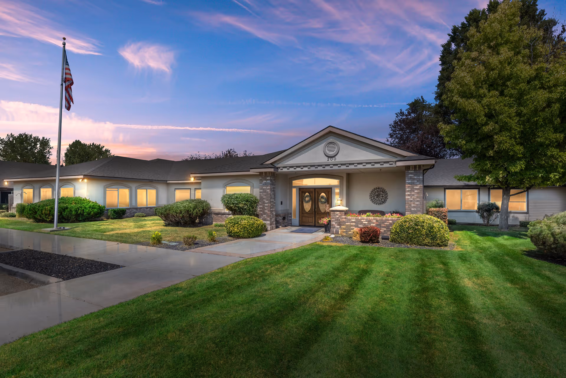 Exterior view of Autumn Wind Assisted Living facility at dusk, showing a single-story building with well-maintained landscaping, green lawn, bushes, trees, a flagpole with an American flag, and a walkway leading to the main entrance with double doors.