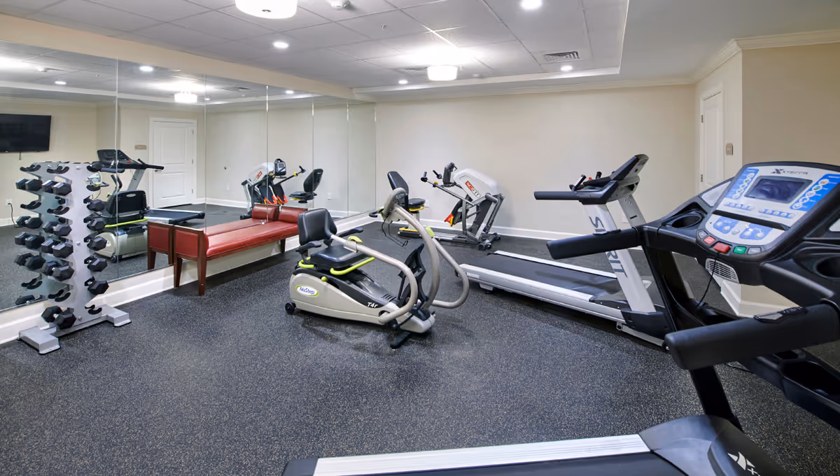 Indoor fitness room with exercise equipment including treadmills, a recumbent bike, a rowing machine, a rack of dumbbells, and a red padded bench. The room has mirrored walls, a wall-mounted TV, and a speckled rubber floor.