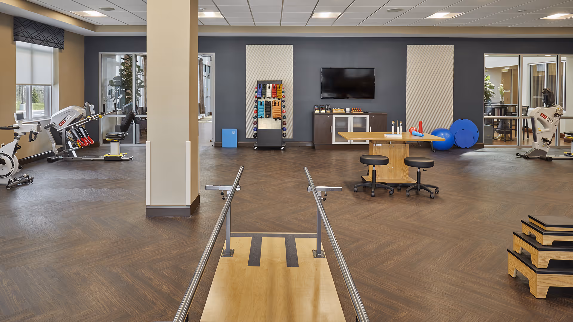 A spacious rehabilitation or physical therapy room with wooden flooring and exercise equipment including stationary bikes, weights, therapy balls, and parallel bars. There is a table with stools in the center, a TV mounted on the wall, and large windows letting in natural light.