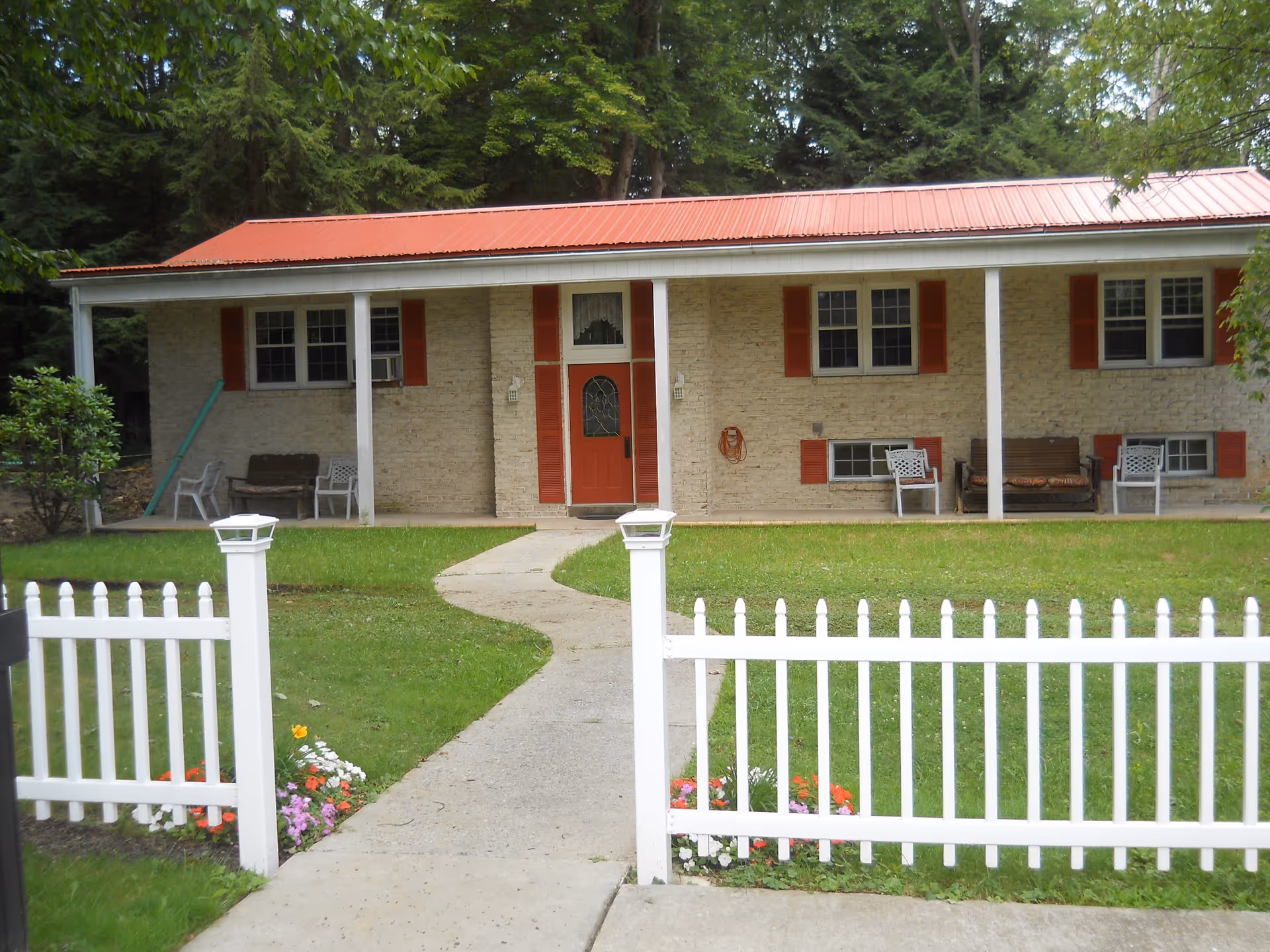 Front exterior view of a single-story brick building with a red metal roof and red shutters. There is a red door in the center with a small porch supported by white columns. The building is surrounded by green grass, a white picket fence, and some colorful flowers near the entrance. Trees are visible in the background.