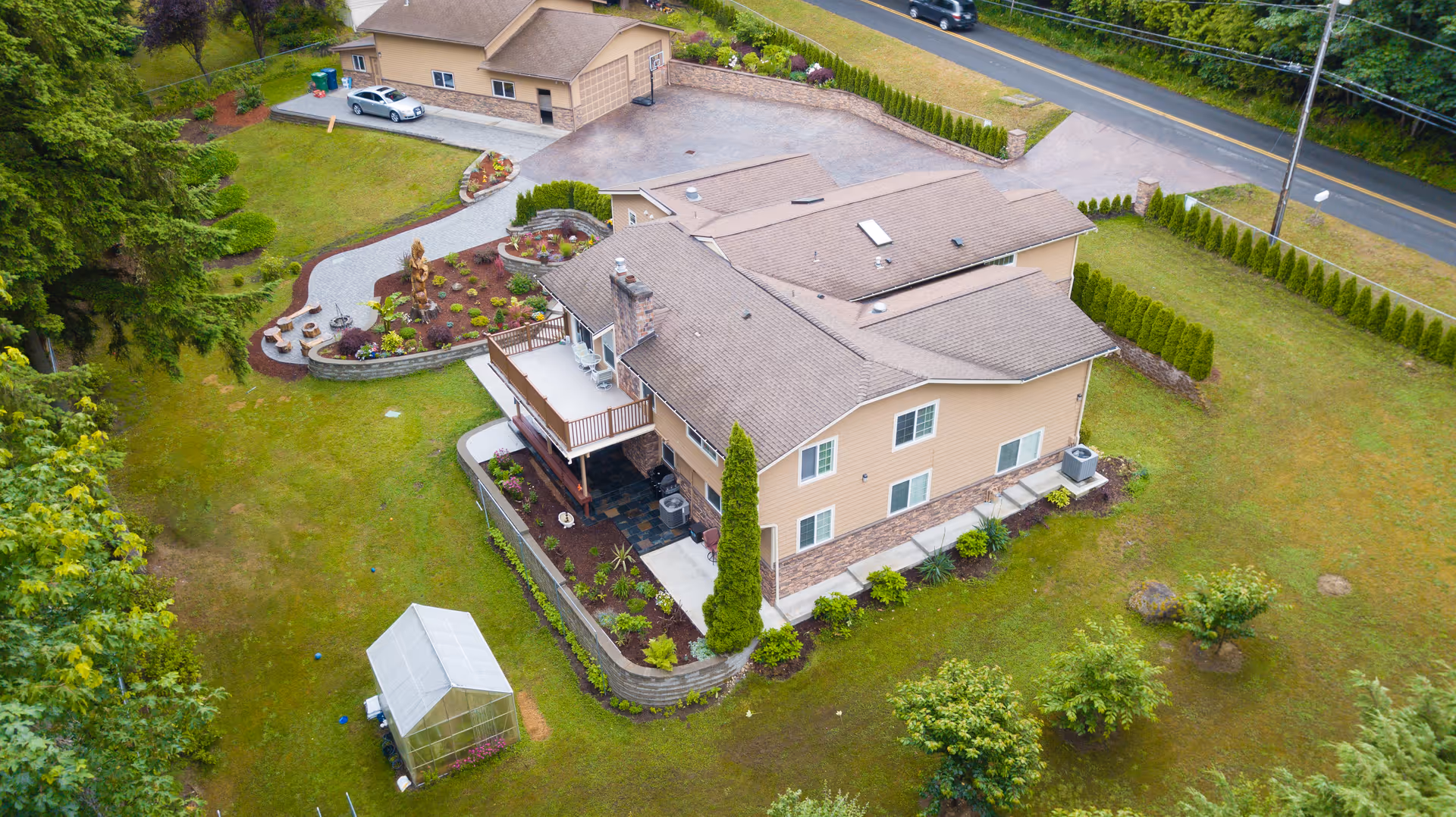 Aerial view of a large beige two-story building with a brown roof, surrounded by a well-maintained lawn and landscaped garden beds. There is a driveway leading to a garage, a small greenhouse in the yard, and a road visible in the background.