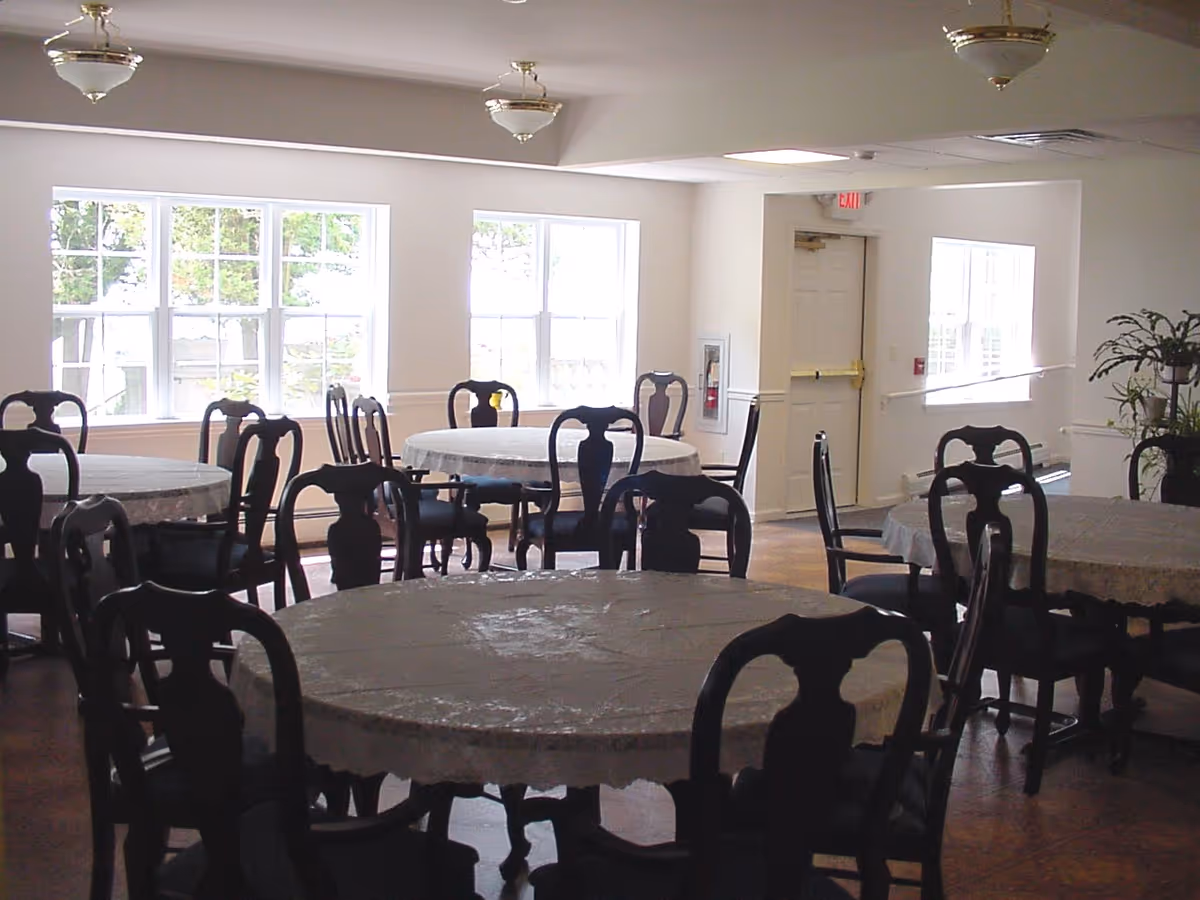 A bright dining room with several round tables covered with white tablecloths and surrounded by dark wooden chairs. Large windows allow natural light to fill the room, and there are ceiling light fixtures. A door and some plants are visible in the background.