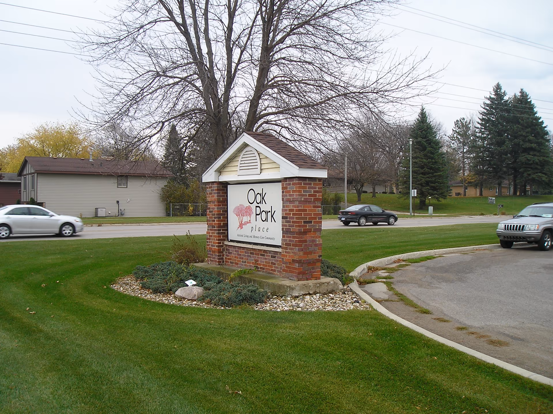 A brick sign with a small roof displaying the name 'Oak Park Place' situated on a grassy area near a road with cars passing by. Trees and houses are visible in the background.