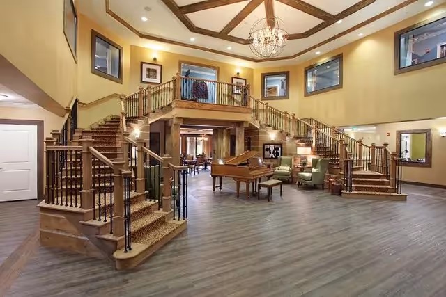 Spacious interior common area with a grand wooden staircase splitting into two directions, a chandelier hanging from the ceiling, a grand piano in the center, and seating arrangements with armchairs and a small table. The walls are painted yellow with framed pictures and windows near the ceiling.