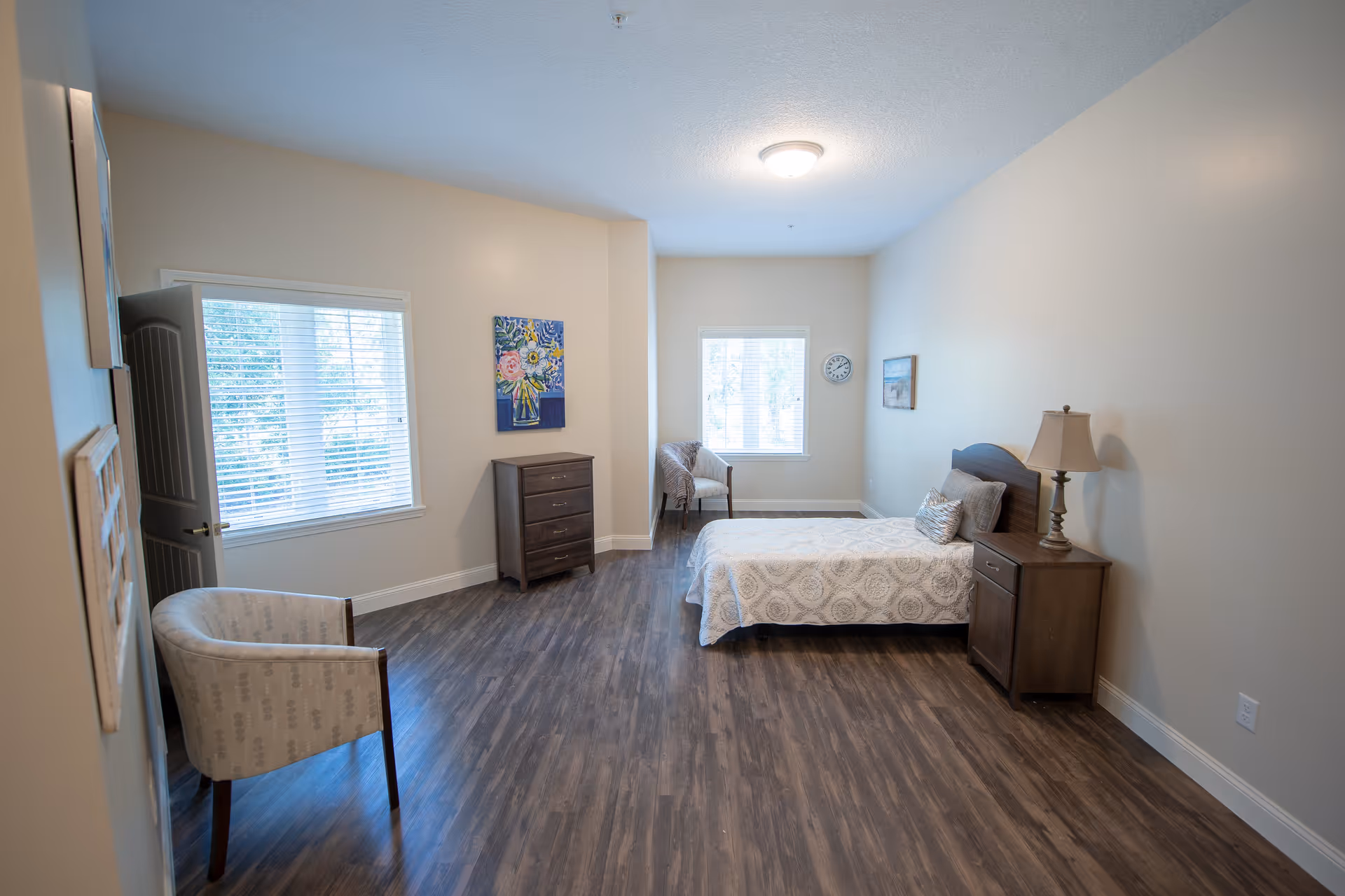 A bright, spacious bedroom with wood flooring, a single bed with patterned bedding, a nightstand with a lamp, two armchairs near windows with blinds, a chest of drawers, and wall decorations including a floral painting and a clock.