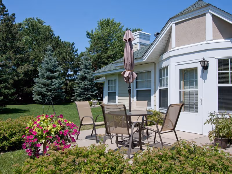 Outdoor patio area at a senior living facility with a round glass table, four mesh chairs, and a closed umbrella. The patio is surrounded by green bushes and flowering plants, with tall trees and a well-maintained lawn in the background. The building has beige siding, white trim, and multiple windows.