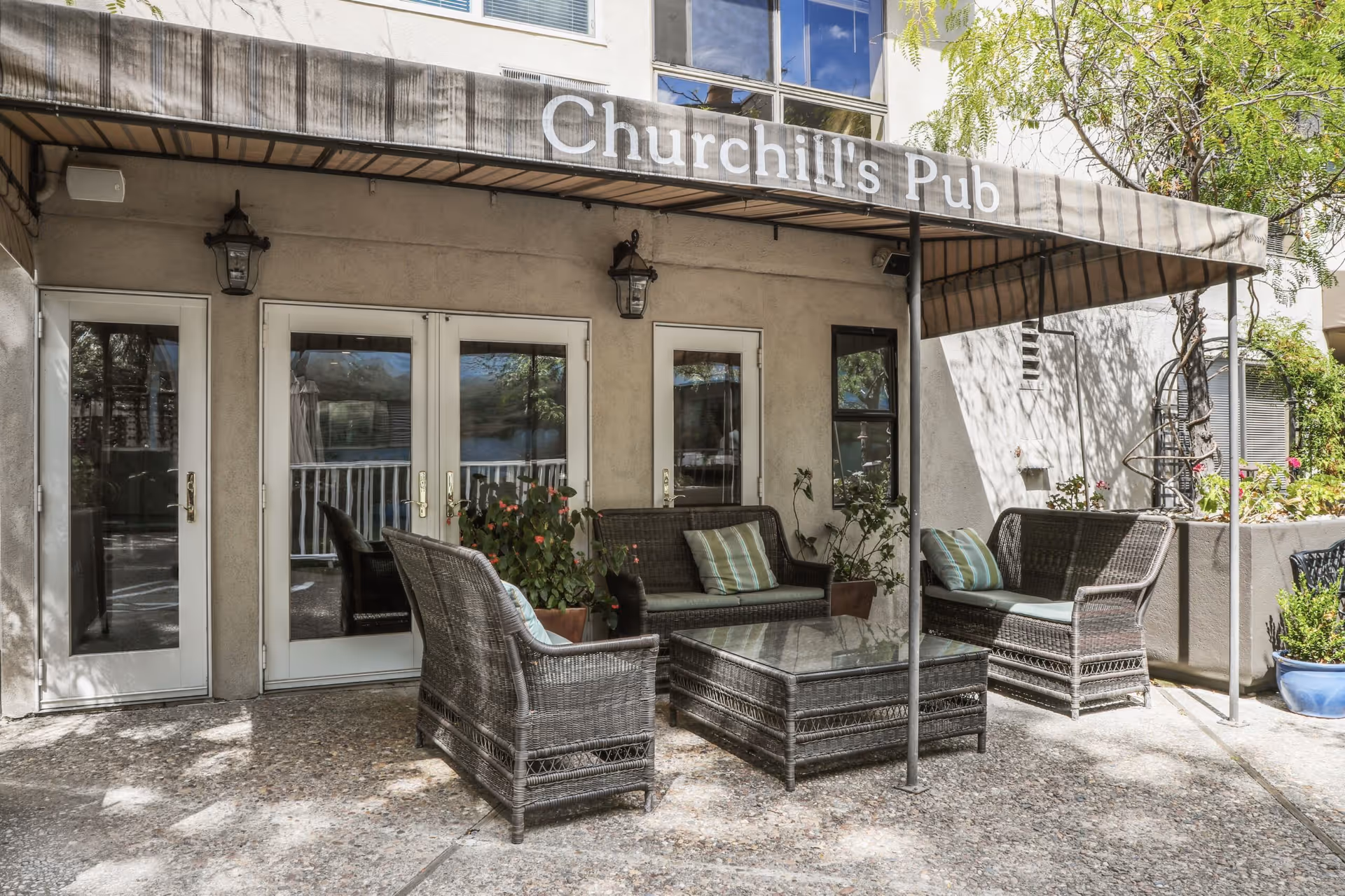 Outdoor patio area with wicker furniture including two armchairs, two loveseats with cushions, and a glass-top coffee table. The area is shaded by an awning labeled 'Churchill's Pub'. There are potted plants around the seating area and a building wall with three glass doors and a window behind the furniture.