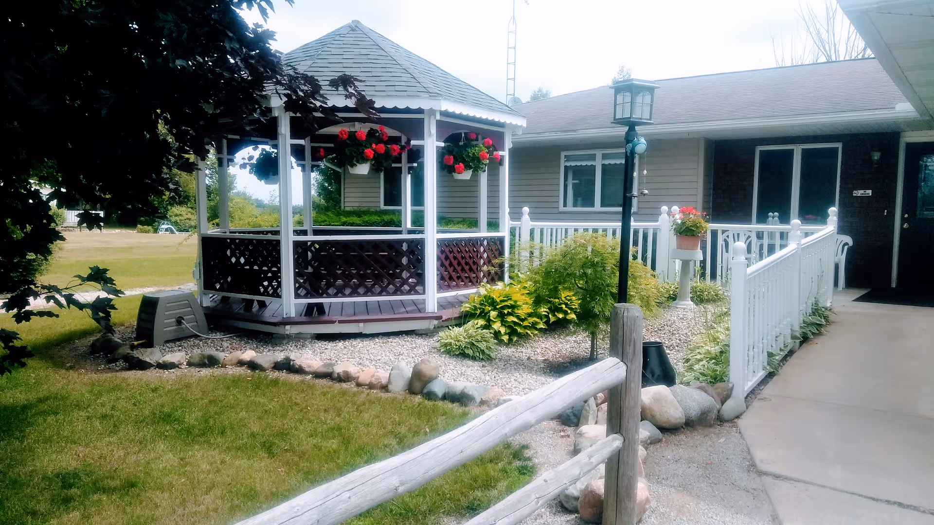 Outdoor view of a senior living facility featuring a white gazebo with hanging flower baskets, a garden area with various plants and shrubs, a white railing along a concrete pathway, and part of the building exterior with windows and a door.