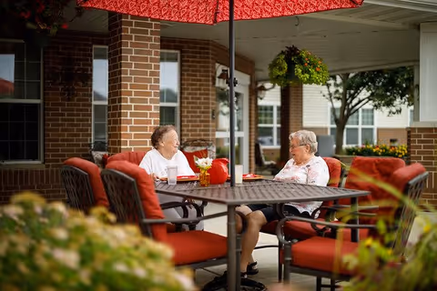 Two elderly women sitting and talking at a round outdoor table with red cushioned chairs under a large red umbrella on a covered patio area of a brick building, with hanging plants and greenery around.