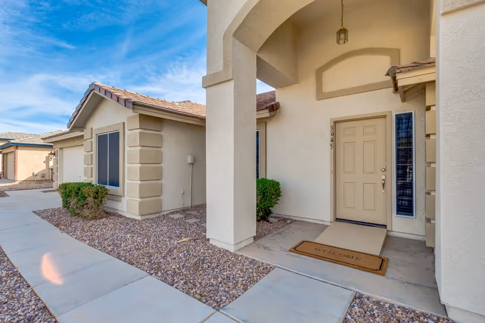 Exterior view of a beige stucco house entrance with a covered porch, a tan front door with the number 3645, a welcome mat, and small bushes along a gravel landscape under a blue sky.
