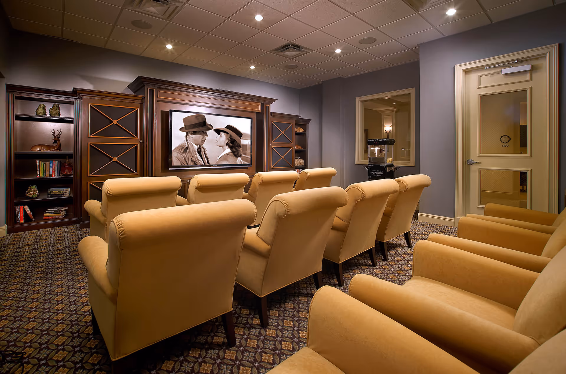 A cozy movie theater room with two rows of beige upholstered armchairs facing a large screen showing a black-and-white film. The room has patterned carpet, built-in wooden shelves with decorative items and books, and a popcorn machine near a door with a window.