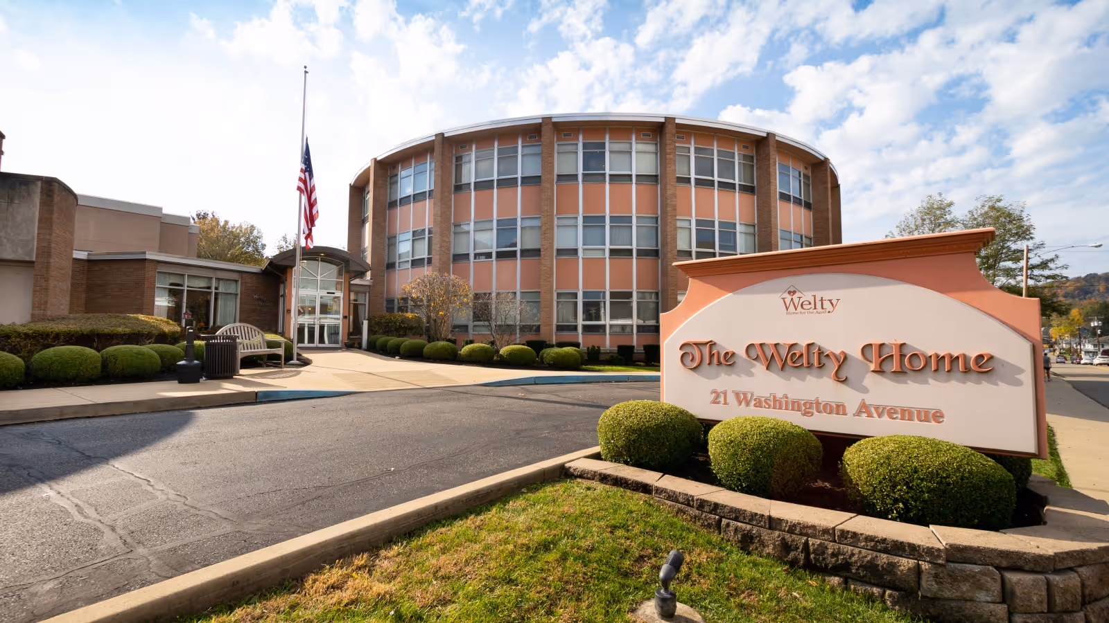 Exterior view of The Welty Home, a senior living facility with a curved three-story building, an American flag at half-mast, a bench, and neatly trimmed bushes. A large sign in the foreground reads 'The Welty Home, 21 Washington Avenue.'