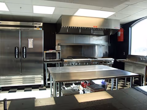 Commercial kitchen with stainless steel appliances including a large refrigerator, microwave, stove with an overhead vent hood, and a stainless steel work table in the center. The floor has a black and white checkered pattern and there is a large window on the right side.