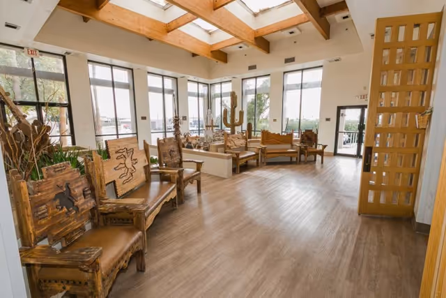 A spacious, well-lit common area with large windows and skylights. The room features rustic wooden chairs and benches with carved designs, arranged along the walls. There are plants and decorative elements, including a large cactus sculpture, contributing to a warm and inviting atmosphere.