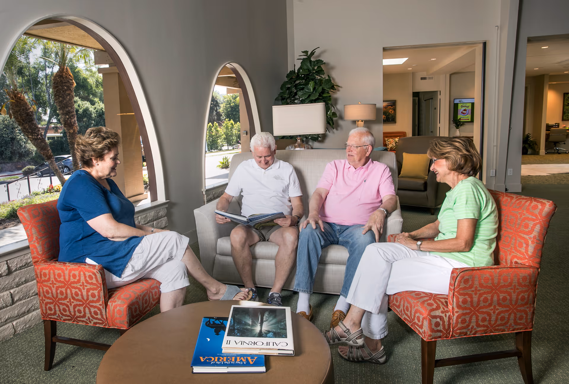 Four elderly people sitting in a living room area with two orange patterned chairs and a beige sofa. Two men are seated on the sofa, one reading a book, while two women sit on the chairs facing them. There are large arched windows showing outdoor greenery, a round coffee table with books, and a lamp with a plant behind the sofa.