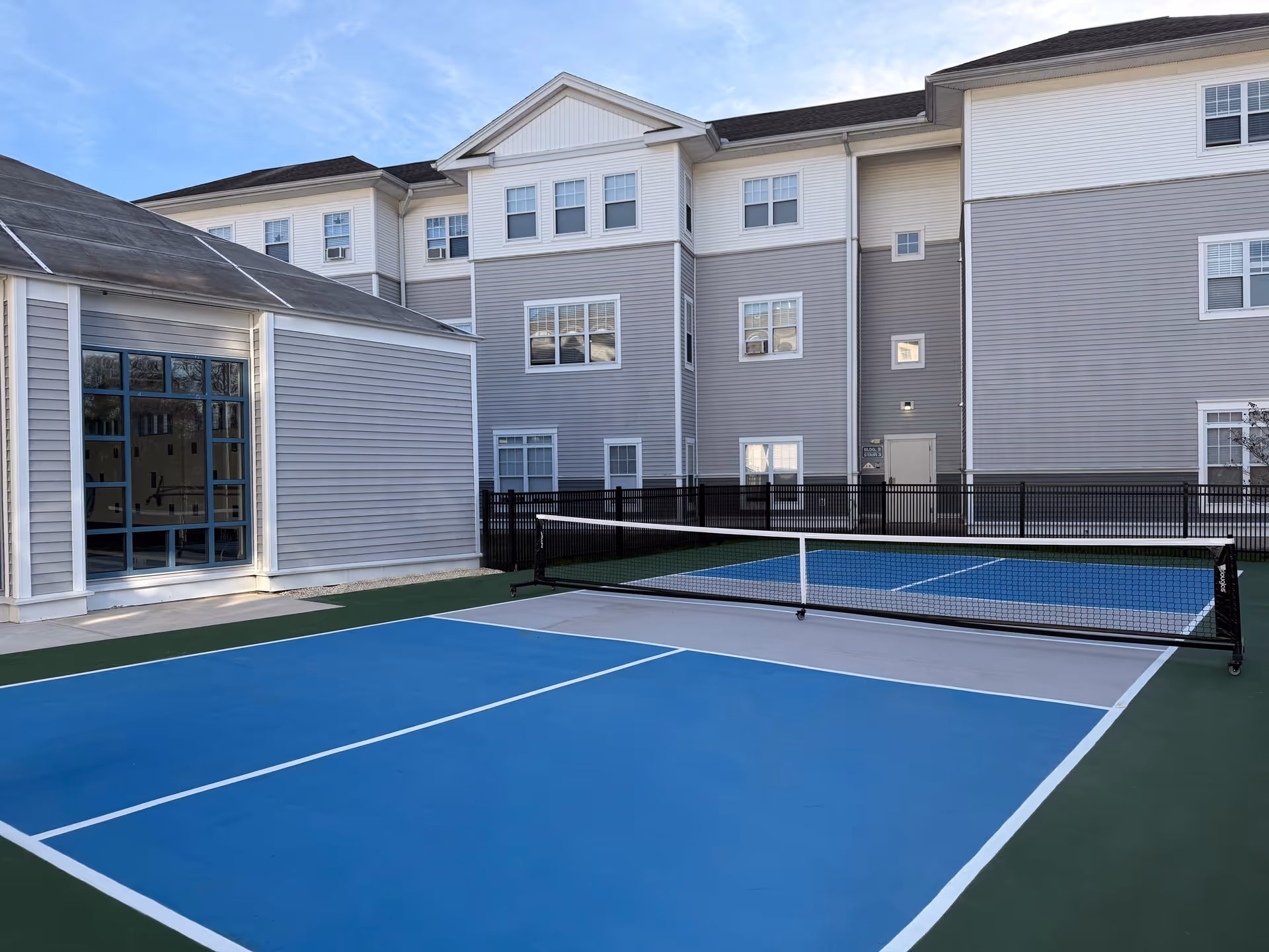 Outdoor view of a pickleball court with a blue and gray playing surface, surrounded by a green border. The court is next to a multi-story building with gray siding and white trim, featuring multiple windows. A black fence encloses the court area, and the sky is clear with some light clouds.
