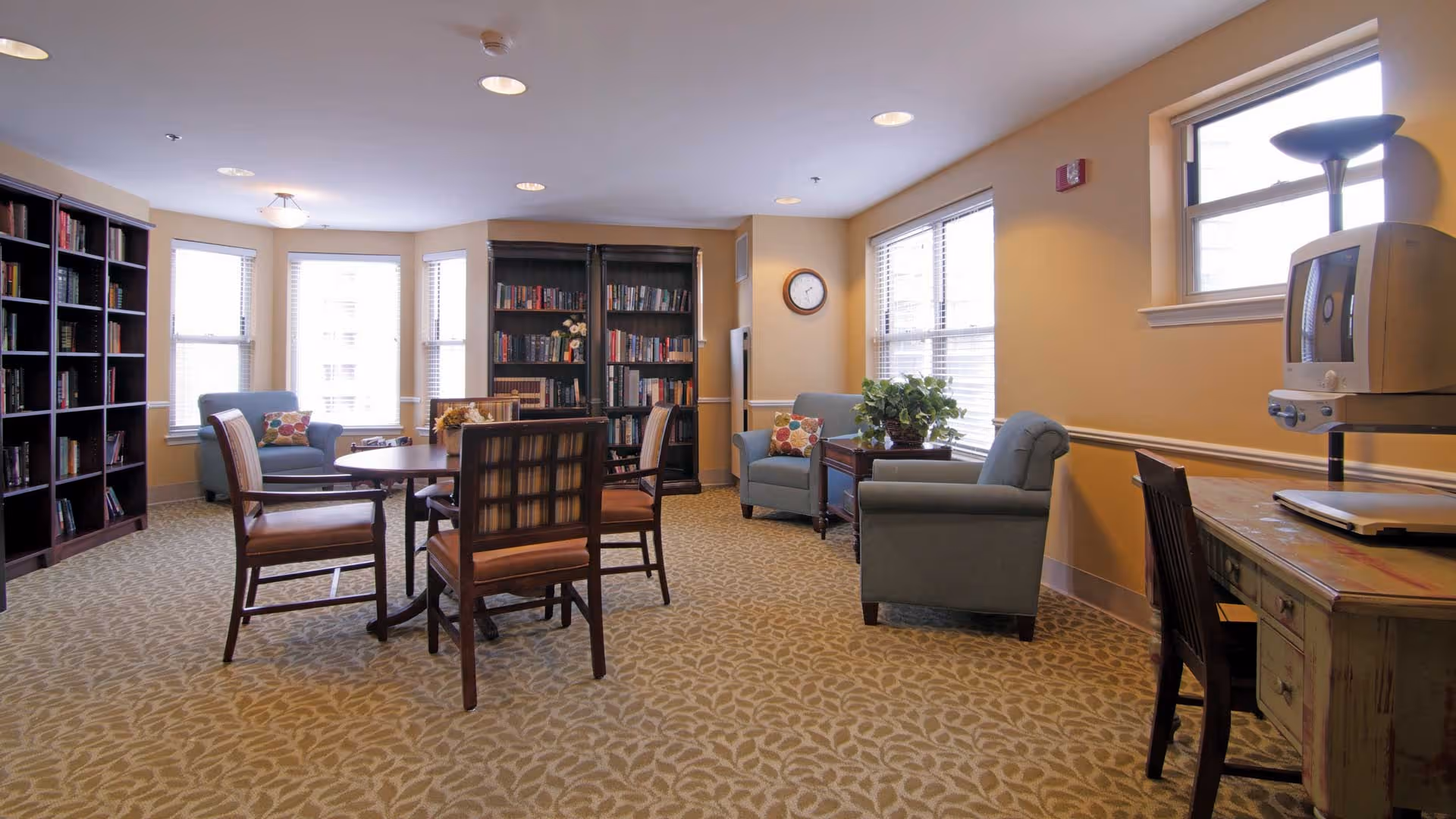 Bright communal lounge with bookshelves, a round table and chairs, upholstered armchairs, and a desk by windows.