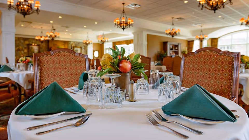 A dining table set for a meal in an elegant dining room with green folded napkins, multiple upside-down glasses, silverware, and a centerpiece with flowers and a red apple. The room features ornate chairs, chandeliers, and large windows allowing natural light.