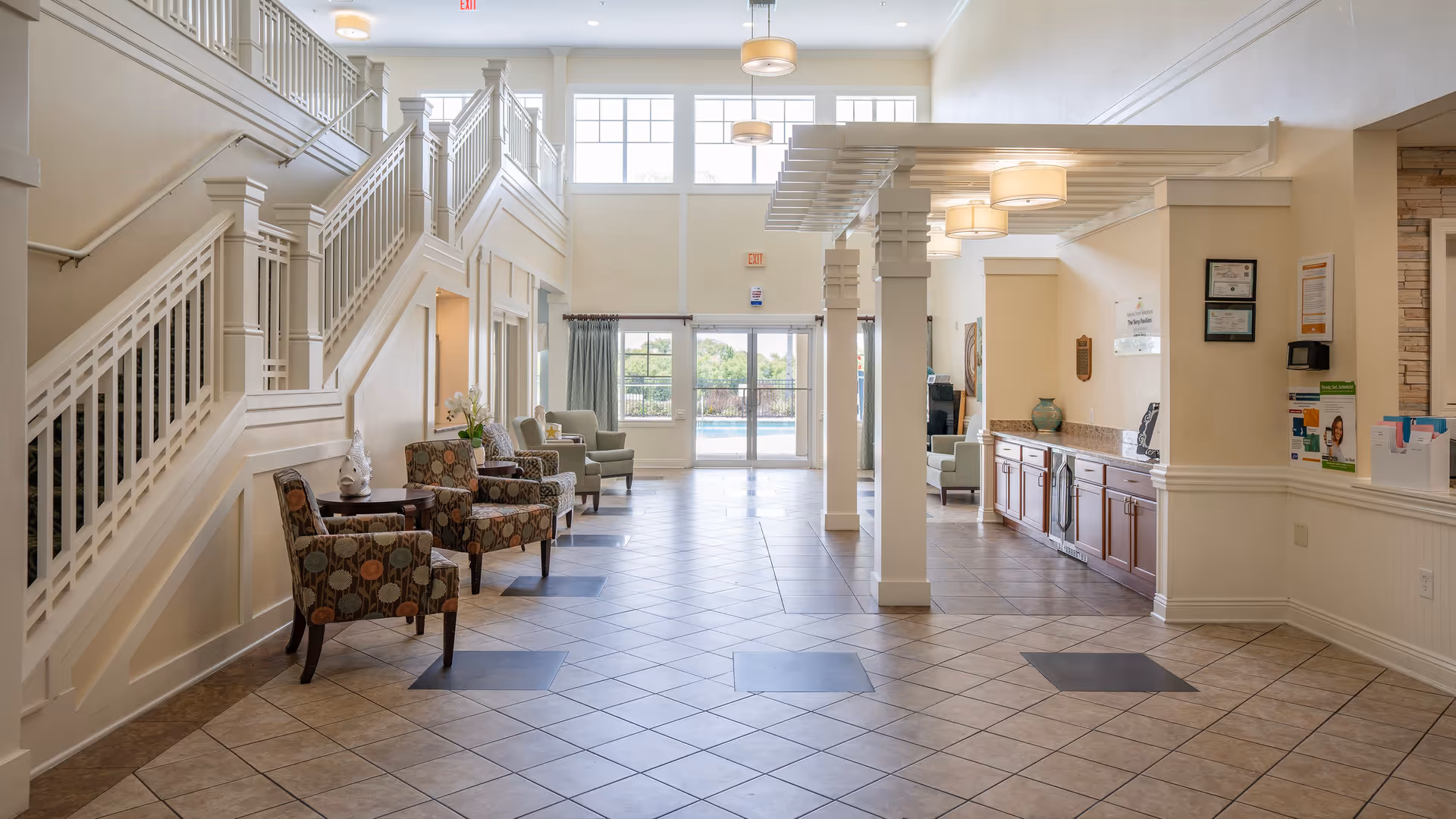 A spacious and well-lit interior common area with tiled floors, patterned armchairs arranged along the left wall, and a staircase with white railings. Large windows and glass doors at the far end allow natural light to fill the space. On the right side, there is a countertop with cabinets underneath and some framed certificates and informational posters on the wall.