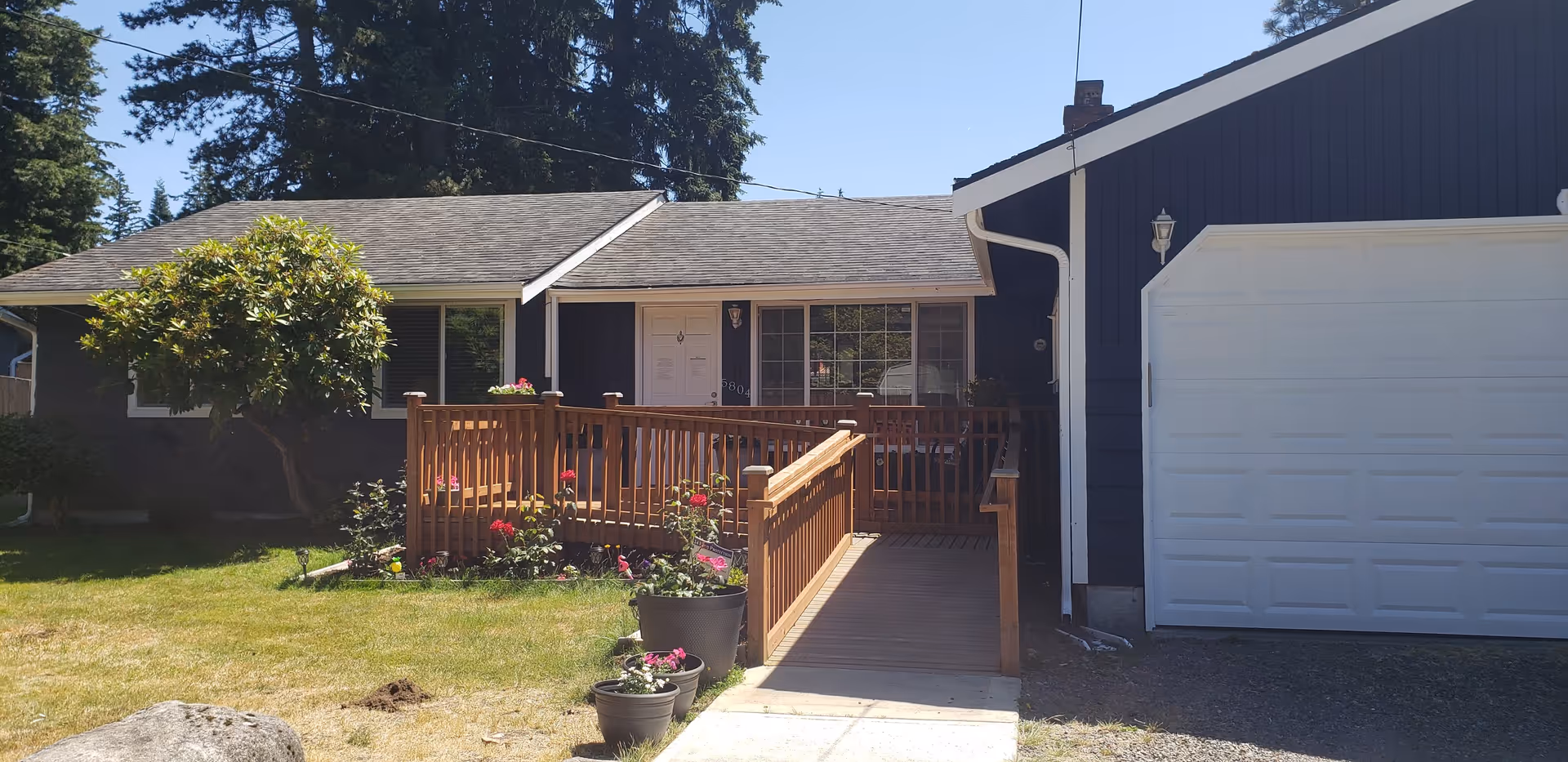 Single-story dark blue house front with a wooden ramp and railing leading to the entrance, an attached white garage, and potted plants in the lawn.