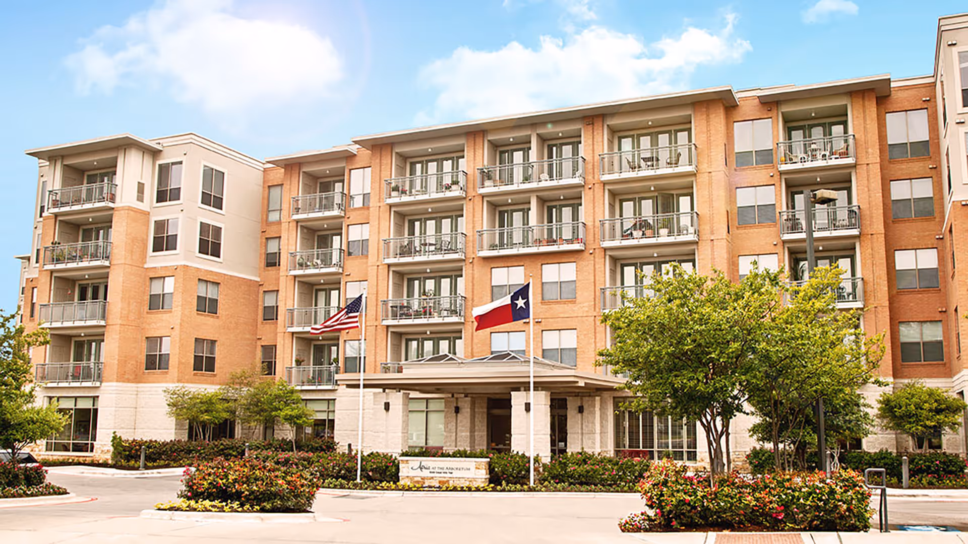 Exterior view of a multi-story senior living facility building with balconies, landscaped greenery, and two flagpoles displaying the American and Texas flags in front of the entrance.