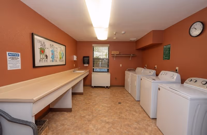 Laundry room with four white washing machines and dryers lined up on the right side. On the left side, there is a long folding counter with a sink. The walls are painted a warm brown color and decorated with framed artwork and informational signs. A window with blinds is centered on the back wall, and a clock is mounted on the upper right wall.
