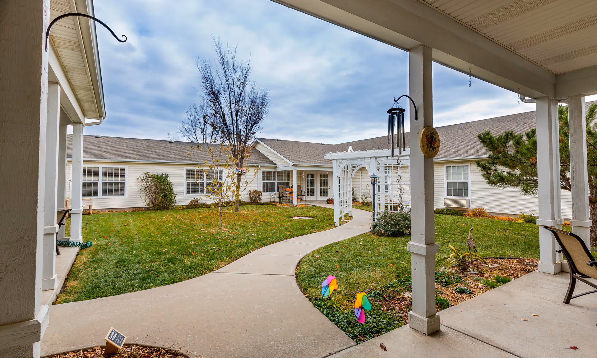 Covered porch framing a central courtyard with a curved walkway, lawn, white pergola, and surrounding single-story building.