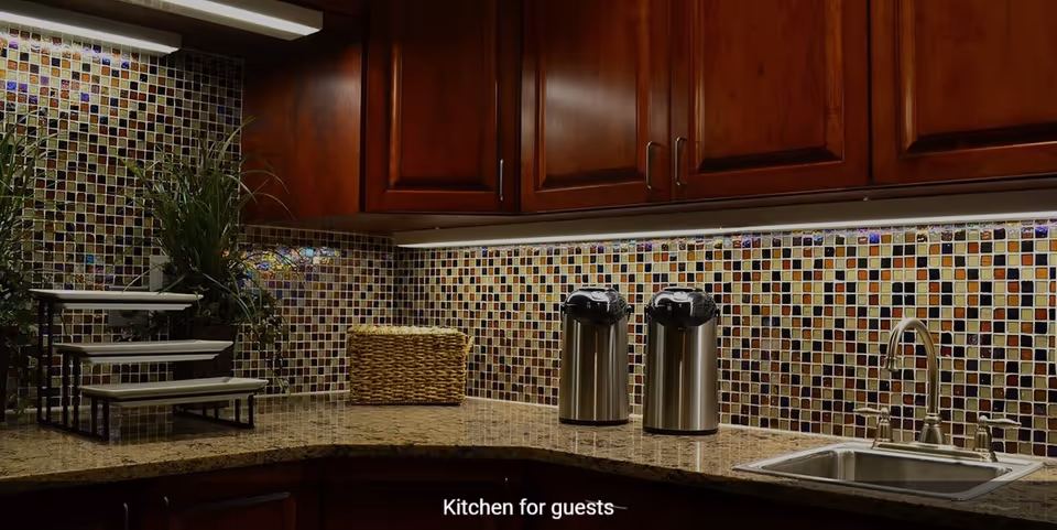 A kitchen countertop with a mosaic tile backsplash, wooden cabinets above, two stainless steel airpots, a woven basket, a small tiered display stand, and a stainless steel sink with a faucet.