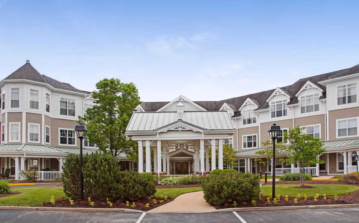 Front entrance of a multi-story senior living building with a covered porte-cochère, landscaped shrubs, and lamp posts.