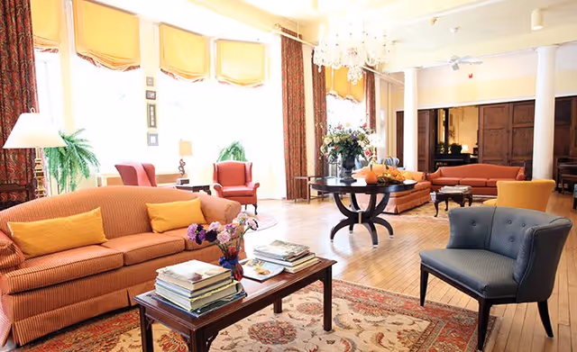 A bright and spacious living room with large windows covered by yellow curtains, multiple upholstered sofas and armchairs in warm colors, a wooden coffee table with books and flowers, a round table with a floral arrangement, hardwood floors, and decorative columns.