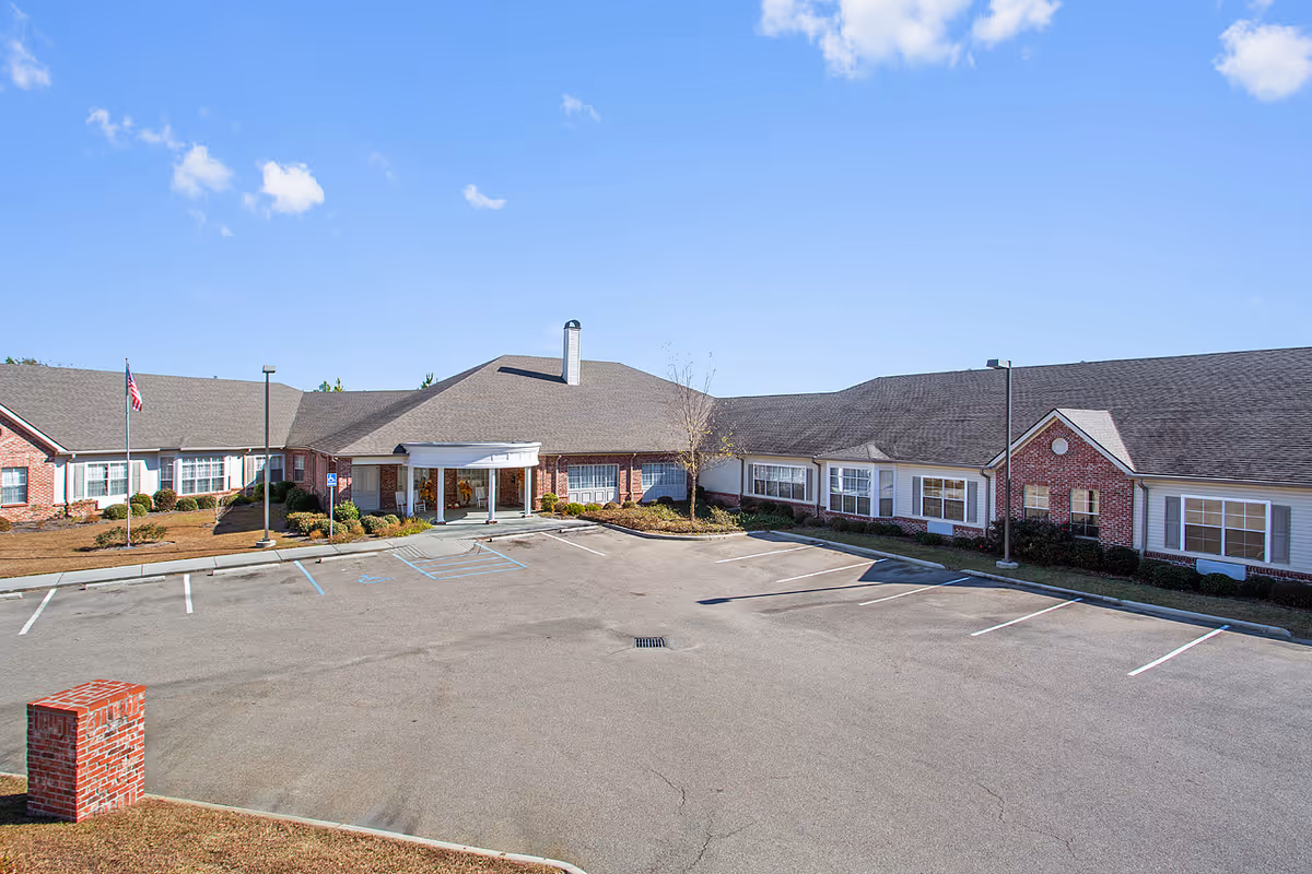 Exterior view of a single-story brick building with a large parking lot in front under a blue sky with scattered clouds. The building has multiple windows and a covered entrance with white columns. There is an American flag on a flagpole near the entrance.