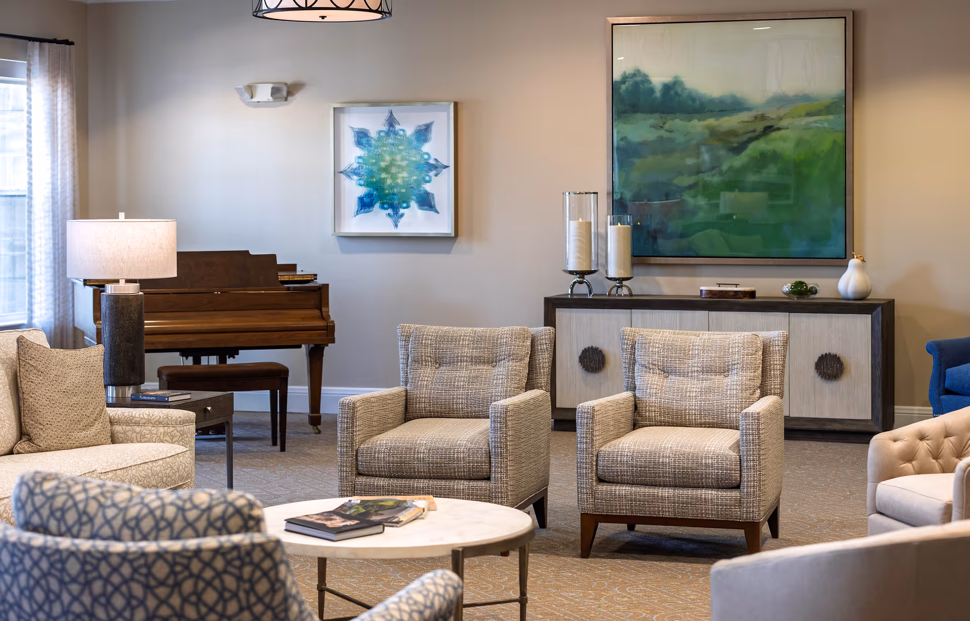 A cozy living room area in a retirement community featuring multiple upholstered armchairs and sofas arranged around a round coffee table with books on it. In the background, there is a wooden piano with a bench, a sideboard with decorative candles and a vase, and two framed abstract paintings on the wall. The room is softly lit with a table lamp and a hanging ceiling light.