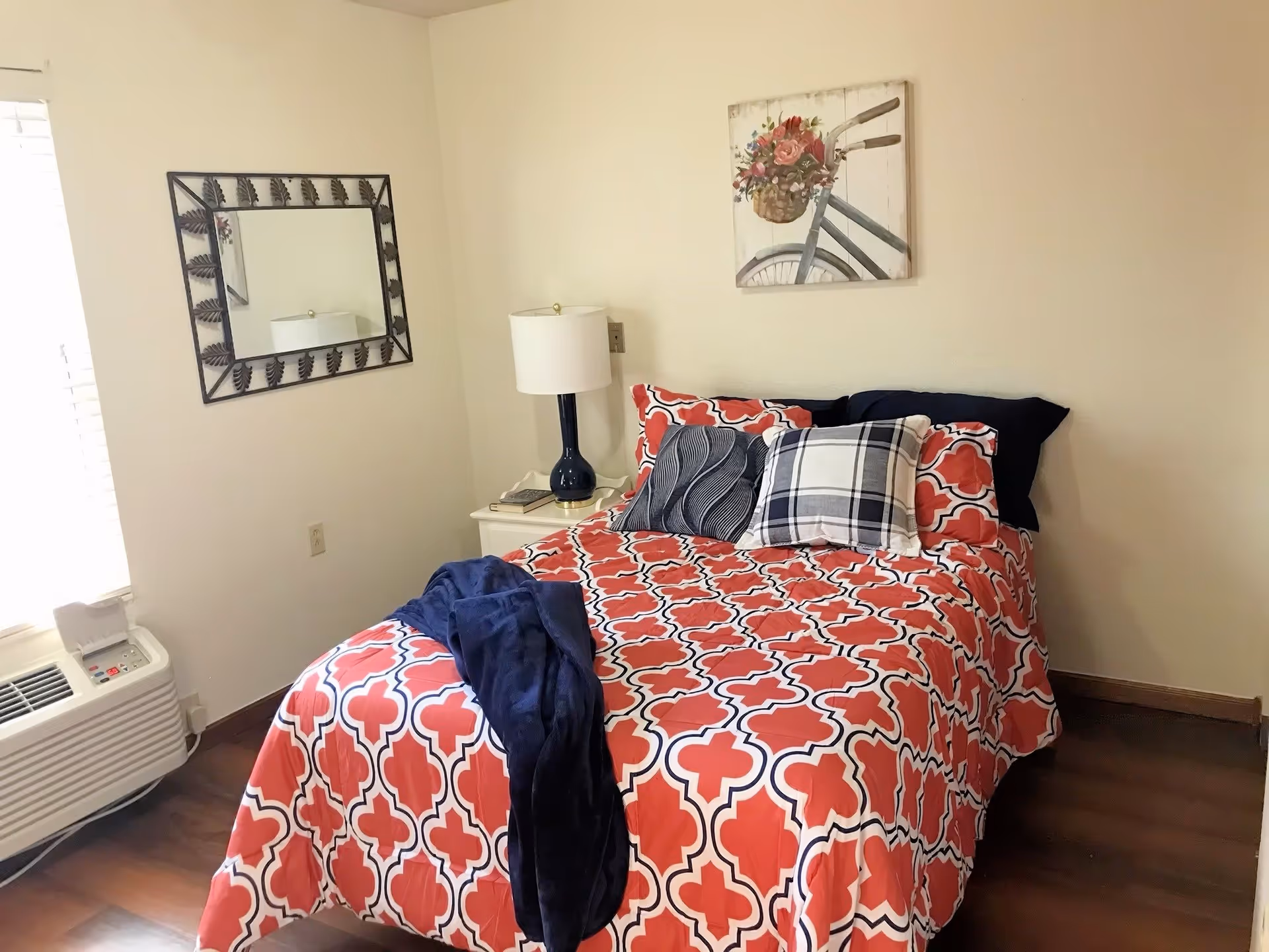 A cozy bedroom with a bed covered in a red and white patterned bedspread, accented with multiple pillows including a plaid one. A navy blue throw blanket is draped over the bed. Next to the bed is a white nightstand with a blue lamp and some books. A decorative mirror with a leaf pattern frame hangs on the wall above the nightstand, and a painting of a bicycle with a basket of flowers is mounted on the wall above the bed. The room has wooden flooring and a window with blinds letting in natural light.