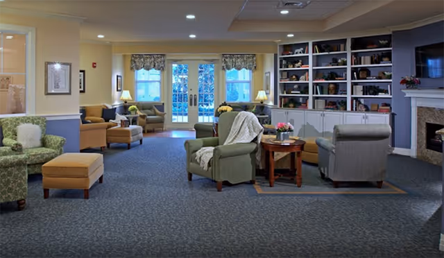 Spacious, well-lit common living room with armchairs, ottomans, side tables, built-in bookshelves and a fireplace facing double doors.