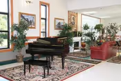 A spacious, well-lit living area featuring a black grand piano on a patterned rug, surrounded by potted plants. The room includes red leather sofas, framed artwork on the walls, large windows, and a fireplace in the background.