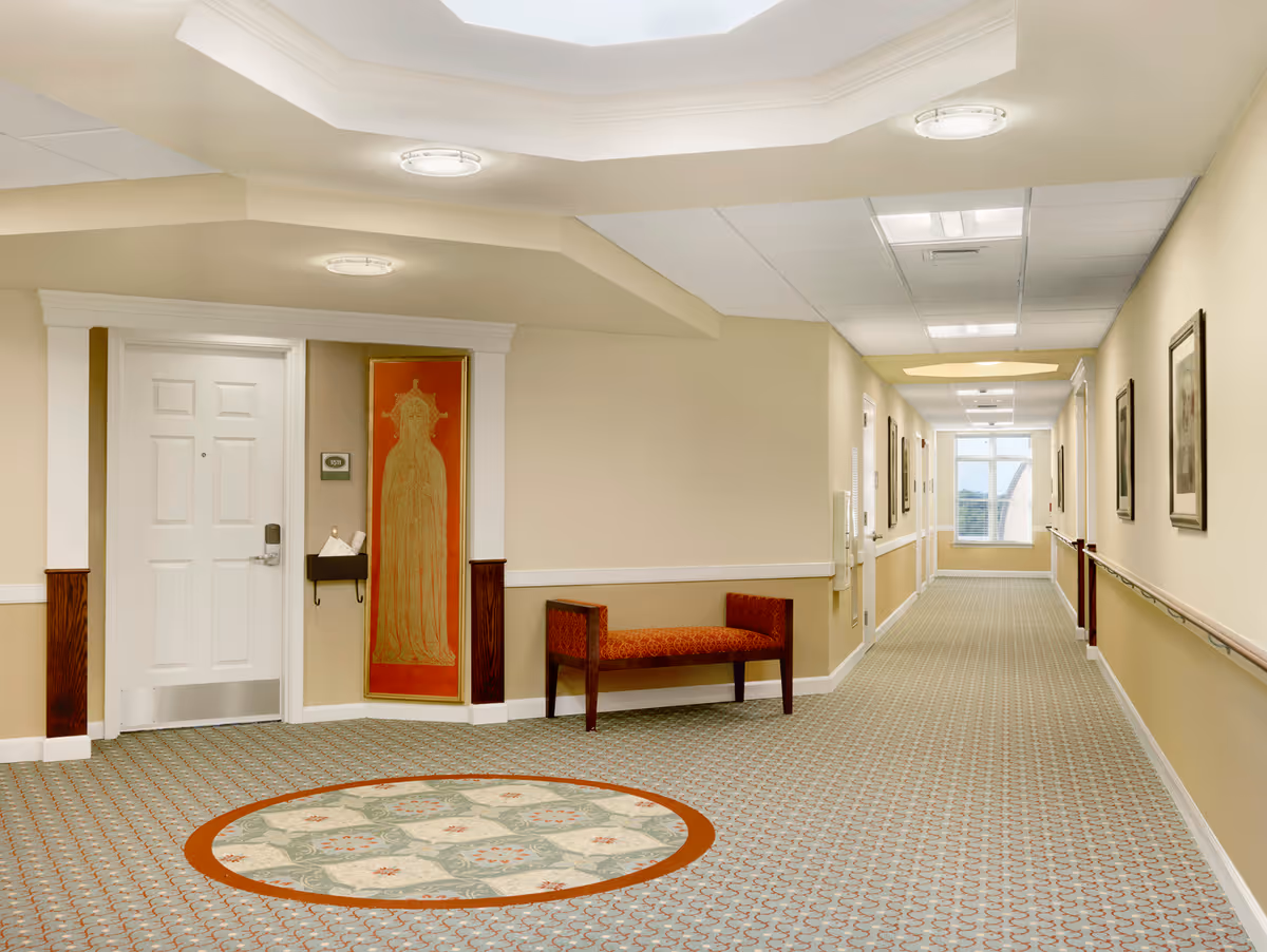 A well-lit hallway in a retirement community with beige walls, patterned carpet, and a white door on the left. There is a red upholstered bench and a decorative wall hanging next to the door. The hallway extends to a window at the far end, with framed pictures on the right wall and handrails along both sides.