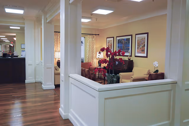 A warm senior living common area showing a reception desk, seating with chairs and a sofa, framed artwork on the wall, and a red floral arrangement.
