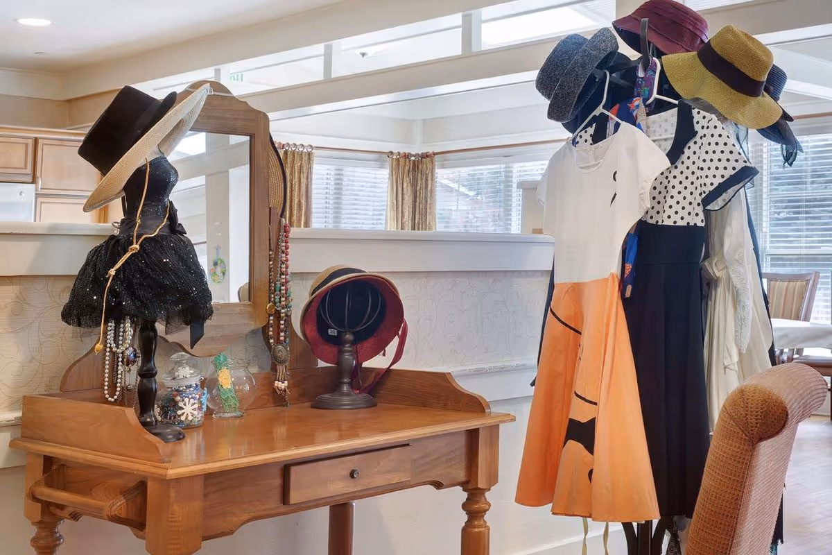 Wooden vanity table and mirror displaying hats, jewelry, and dresses on stands in a bright communal room.