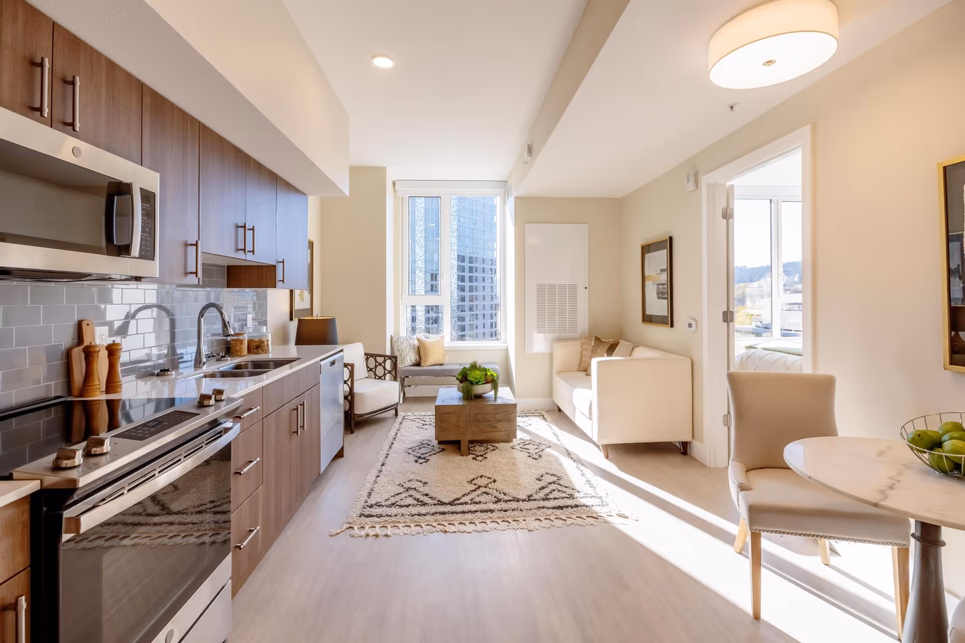 Bright and modern open-concept living space featuring a kitchen with wooden cabinets, stainless steel appliances, and a gray tiled backsplash on the left. The room extends to a cozy sitting area with a beige sofa, armchair, and a wooden coffee table on a patterned rug. Large windows at the back let in natural light and offer a view of tall buildings outside. On the right, there is a small round dining table with two beige chairs and a bowl of green apples. The walls are light-colored with framed artwork and a ceiling light fixture.