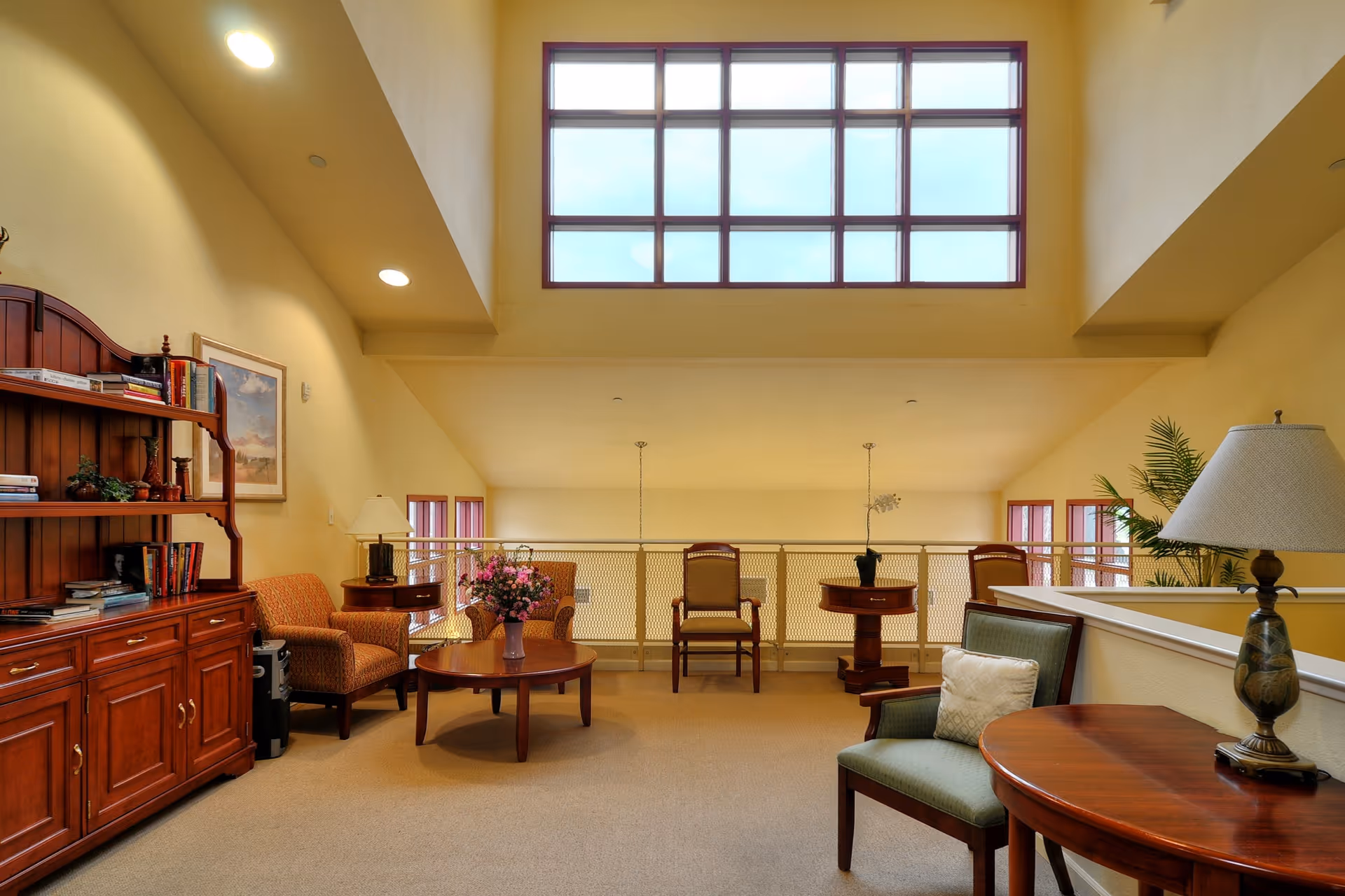 A spacious and well-lit living area with high ceilings and large windows. The room features wooden furniture including a bookshelf filled with books, a round coffee table with a flower vase, several chairs, a side table with a lamp, and a potted plant. The walls are painted a warm yellow color.