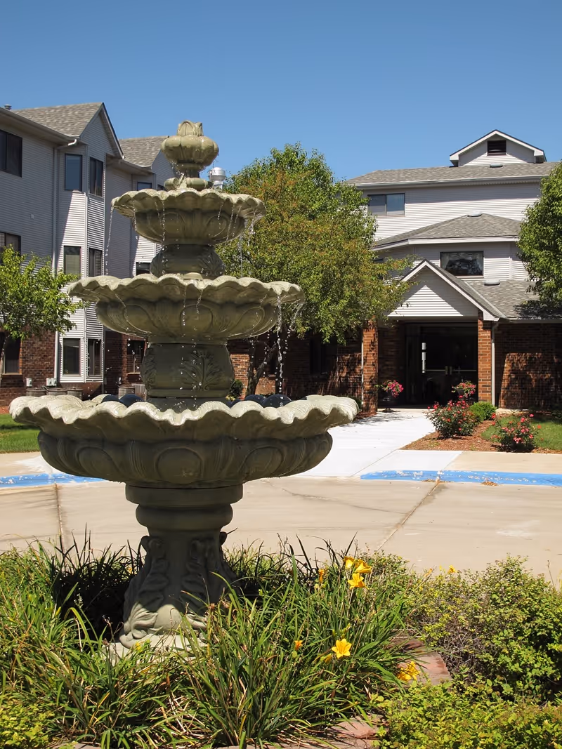 A tiered stone fountain in front of the entrance of a multi-story senior living building with trees and flowers.