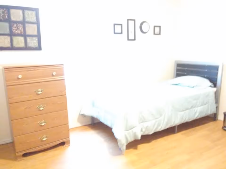 A simple bedroom with a single bed covered in a light-colored comforter and a pillow. Next to the bed is a wooden chest of drawers with four drawers and brass handles. The walls are white and decorated with framed artwork above the bed and chest of drawers. The floor is wooden.