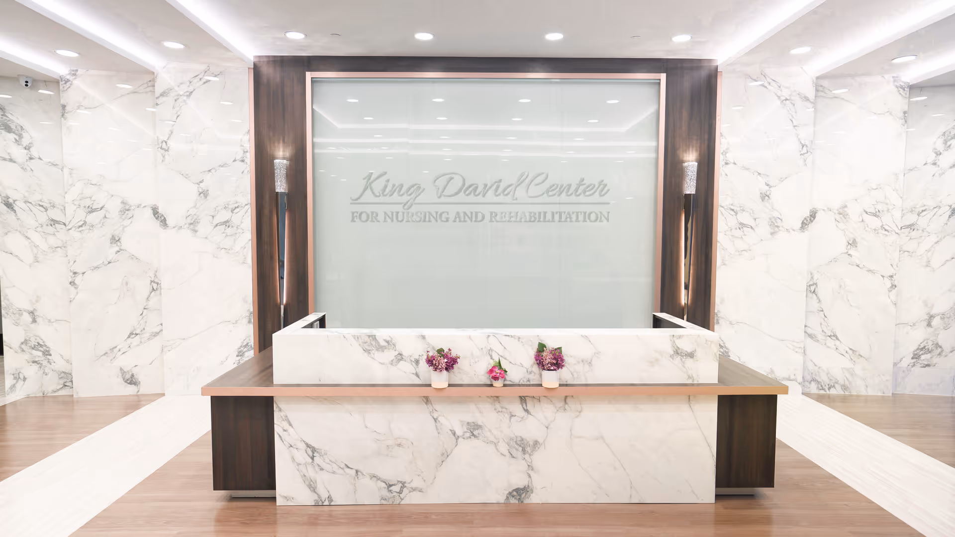 A modern reception desk area with white marble walls and a marble front desk featuring a wooden countertop. Behind the desk is a large frosted glass panel with the text 'King David Center for Nursing and Rehabilitation'. There are two small flower pots with purple and pink flowers on the desk. The floor is a combination of wood and white tiles, and the space is well-lit with ceiling lights.
