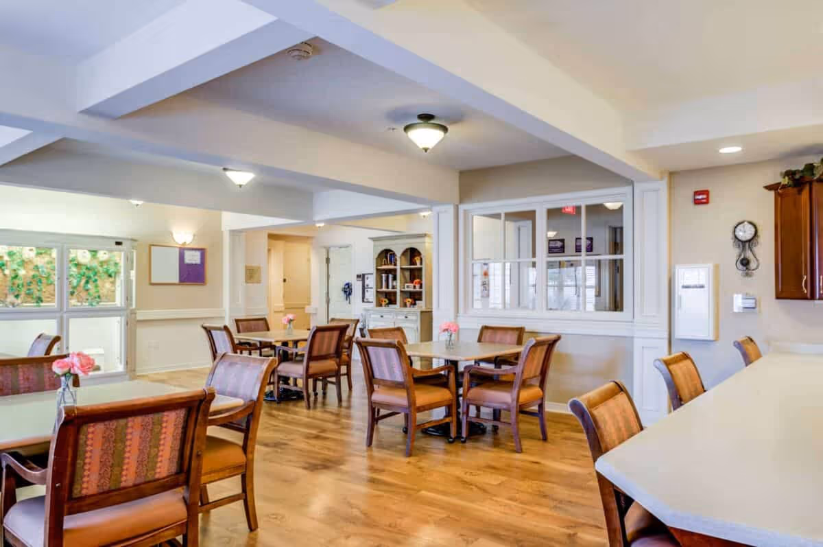 Bright communal dining room with wooden tables and chairs, small vases of flowers, and a service counter in a senior living facility.